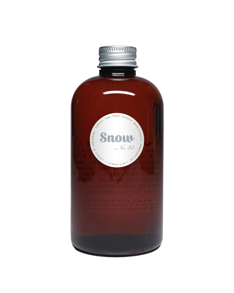 A brown glass bottle with a silver screw cap and round white label reading "Simpatico Snow Diffuser Refill" stands upright against a plain white background, representing the winter scent product from Simpatico.