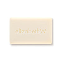 The elizabeth W Botanist's Scent Lab Lilac Bar Soap, rectangular in shape and embossed with the brand name "elizabethW," emits a gentle floral aroma. This creamy, light lilac-colored soap is presented against a plain white background.