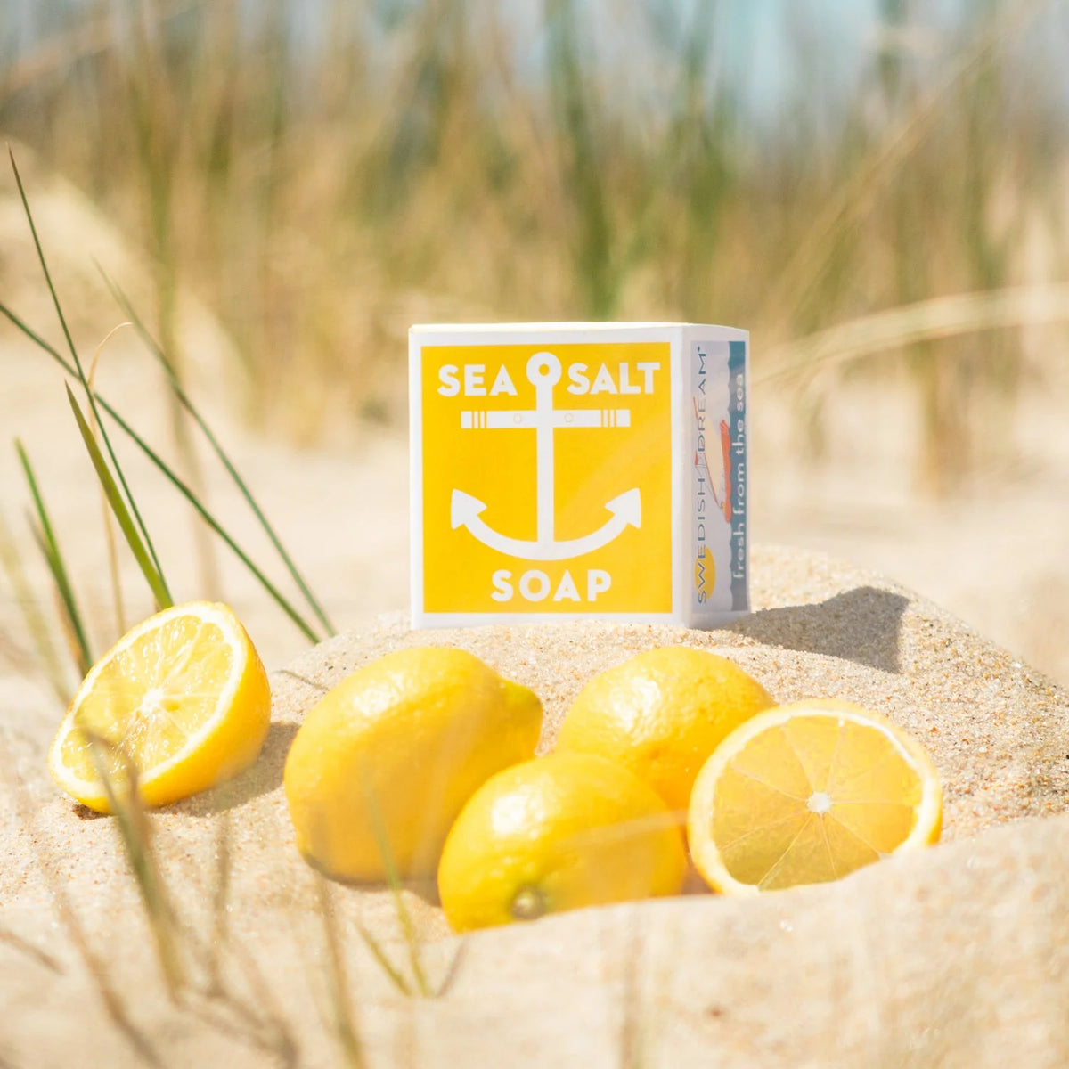 A bar of Swedish Dream Sea Salt Lemon Soap with a yellow anchor design is placed on sandy ground alongside several fresh lemons. The background features beach grasses and blurry ocean waves, evoking a refreshing coastal setting infused with lemon citrus oils.