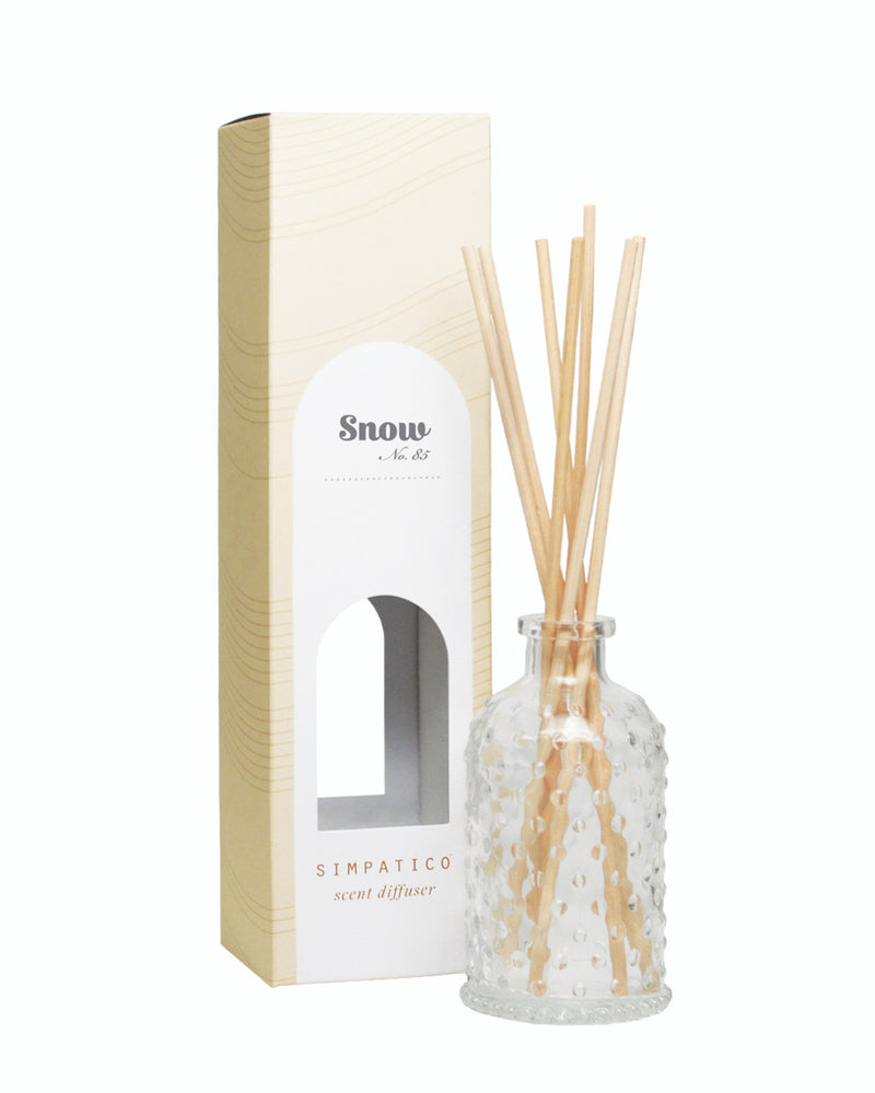 An image of a Simpatico Snow Hobnail Diffuser Kit featuring a clear, decorative glass bottle with several light wood reeds inserted, beside its beige packaging box with a window revealing the.