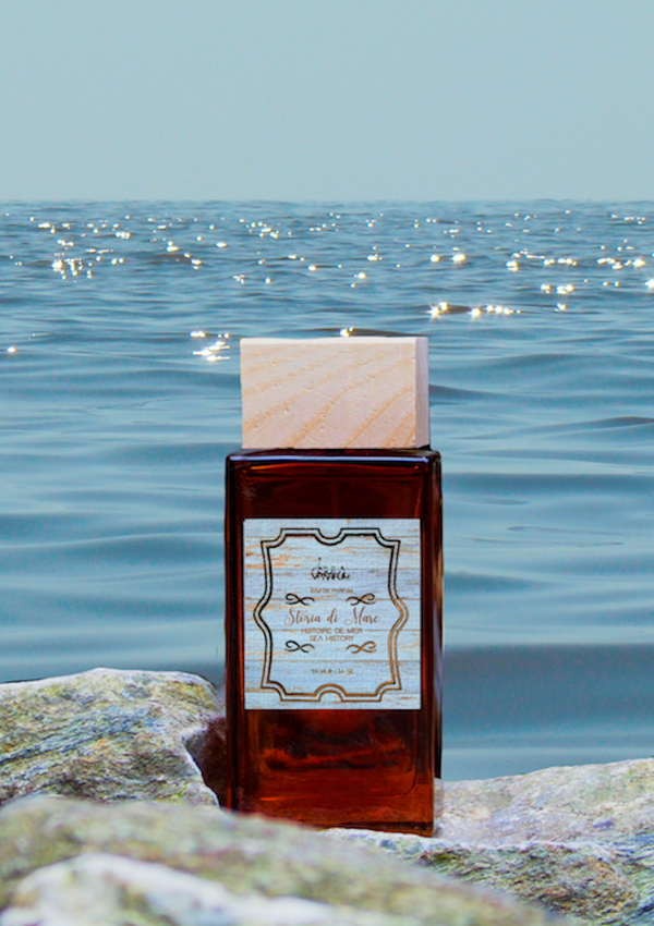 The ISULA parfums Storia di Mare - Sea Story eaux de parfum sits on seaside rocks, its brown glass bottle with a wooden cap reflecting sunlight and blue waves—evoking aquatic freshness and subtle sea salt notes by the shore.