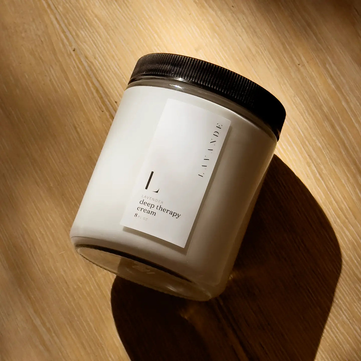 A white jar with a black lid, labeled "Lavande - Deep Therapy Cream 8oz" by Lavande, enriched with lavender essential oil, rests on a sunlit wooden surface and casts a gentle shadow.