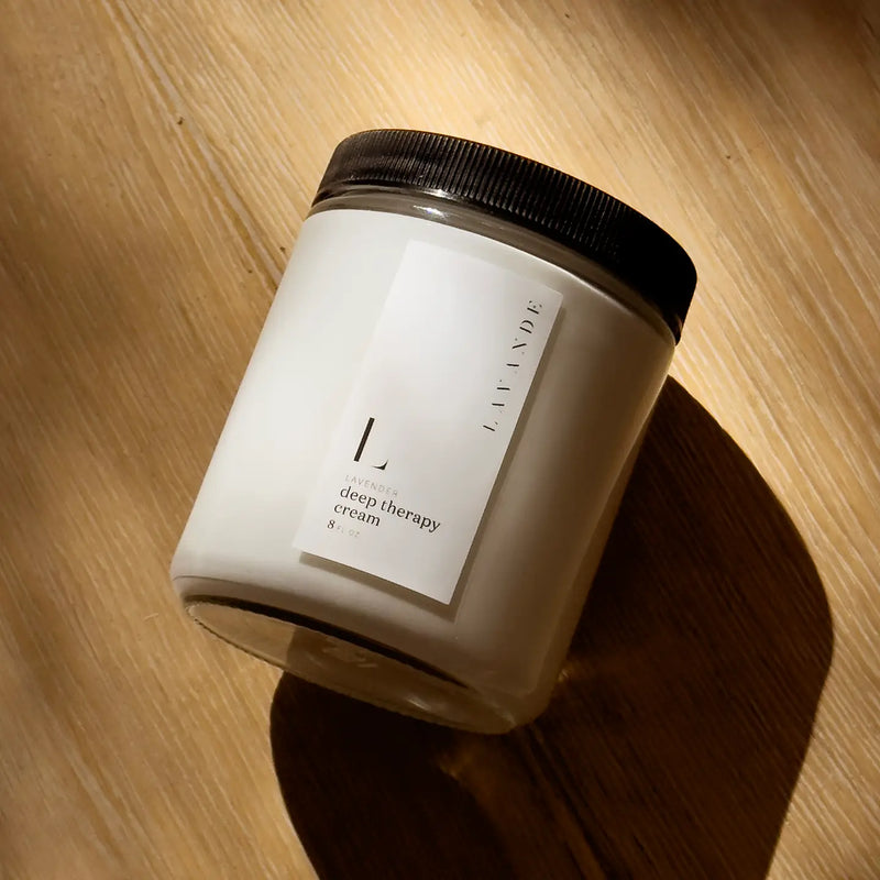 A white jar with a black lid, labeled "Lavande - Deep Therapy Cream 8oz" by Lavande, enriched with lavender essential oil, rests on a sunlit wooden surface and casts a gentle shadow.