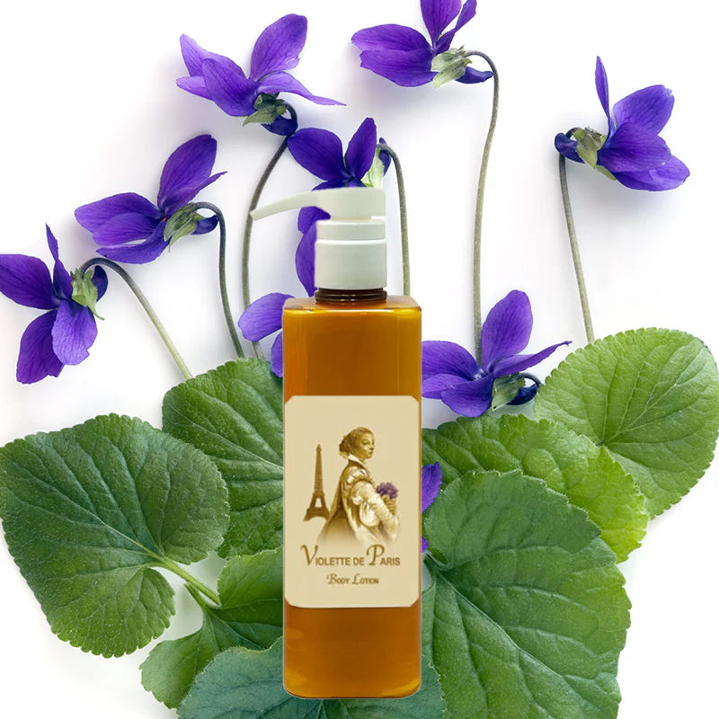 A bottle of La Bouquetiere Violette de Paris Body Lotion sits before green leaves and purple violets, promising youthful elasticity, all set against a white background.