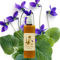 A bottle of La Bouquetiere Violette de Paris Body Lotion sits before green leaves and purple violets, promising youthful elasticity, all set against a white background.