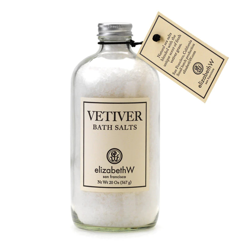 A glass bottle of elizabeth W Vetiver Bath Salts with a beige label and a hanging tag describing the soothing bath product, isolated on a white background.