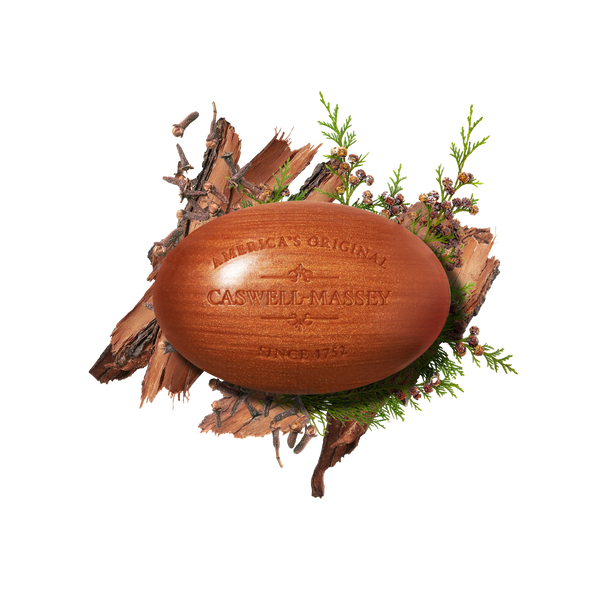 A brown oval Caswell-Massey Sandalwood Woodgrain Bar Soap engraved with "America's Original Caswell-Massey Since 1752," rests on bark and greenery with purple berries on a white background.