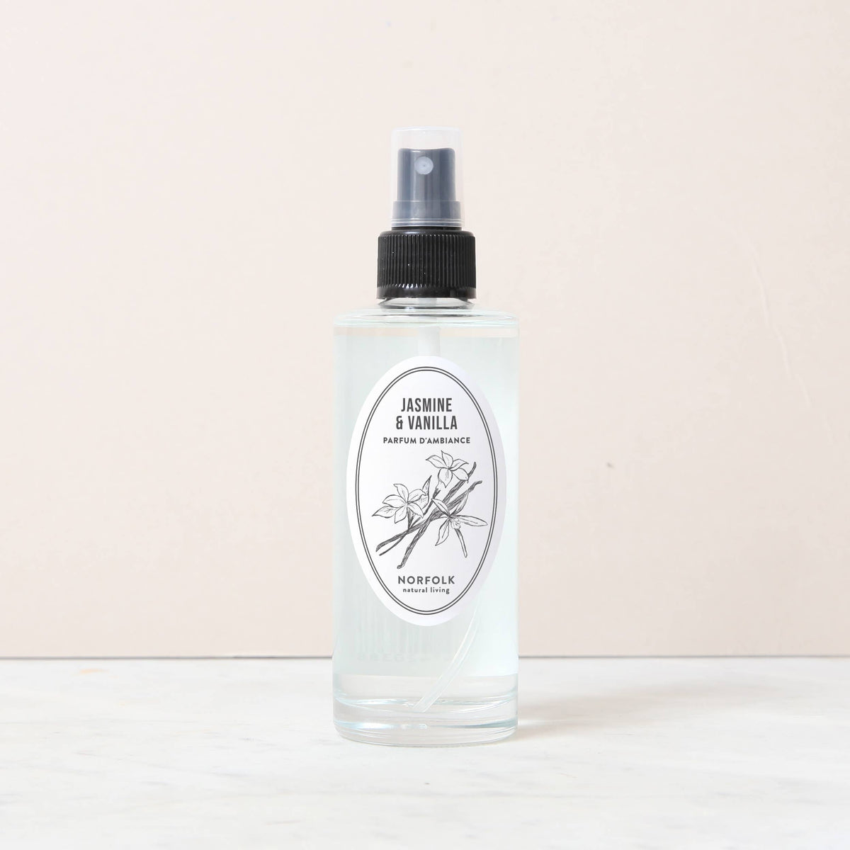 A clear spray bottle of Norfolk Natural Living Jasmine & Vanilla Room and Mood Mist sits on a white surface, its label featuring a botanical illustration that evokes the fresh jasmine and vanilla scent.