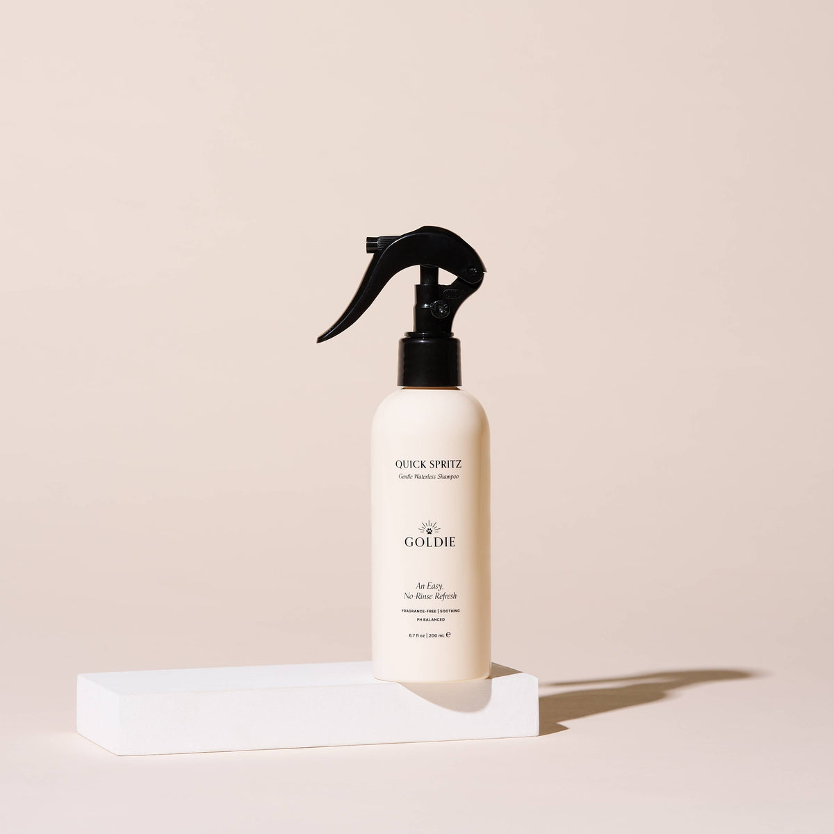A beige spray bottle of Goldie - Quick Spritz, a fragrance-free dog shampoo by Goldie, stands upright on a white rectangular block with a plain light beige background.
