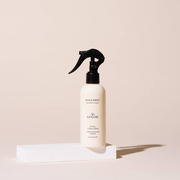 A beige spray bottle of Goldie - Quick Spritz, a fragrance-free dog shampoo by Goldie, stands upright on a white rectangular block with a plain light beige background.