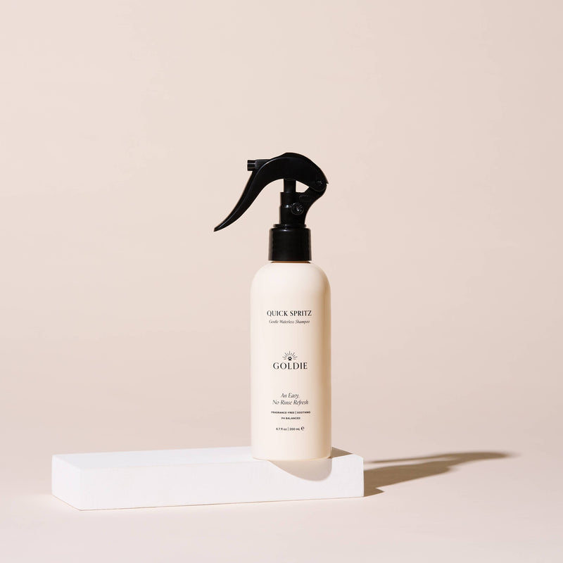 A beige spray bottle of Goldie - Quick Spritz, a fragrance-free dog shampoo by Goldie, stands upright on a white rectangular block with a plain light beige background.