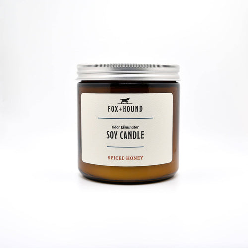 The Fox + Hound Odor Eliminator Soy Candle in Spiced Honey features a brown jar with a silver lid, offering a cozy atmosphere and soothing scent. Its elegant design on a white background invites warmth and comfort.