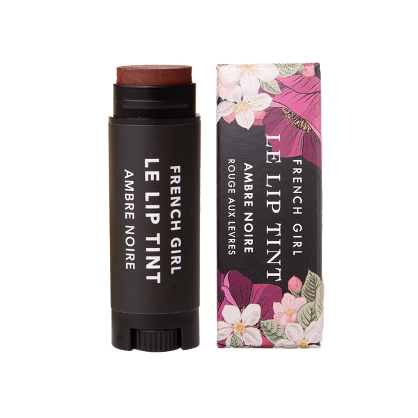 A tube of FRENCH GIRL Tinted Lip Balm in Ambre Noire stands by its floral box, both with white text on black. This vegan balm by FRENCH GIRL hydrates lips with a dark reddish-brown tint for a natural look.