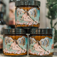 A trio of jars, showcasing labels adorned with a vintage-style illustration of a woman with butterfly wings amidst roses, suggests the nourishing and healing properties of Barefoot Venus The Vanilla Effect Instant Hand Repair Balm. These jars feature black lids and are set against a backdrop that includes hints of greenery and blurred bottles.