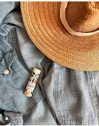 A Stewart & Claire Negroni Lip Balm in a paper tube rests atop a straw hat and denim shirt, evoking a casual summer vibe and showcasing this eco-friendly lip care essential for all your adventures.