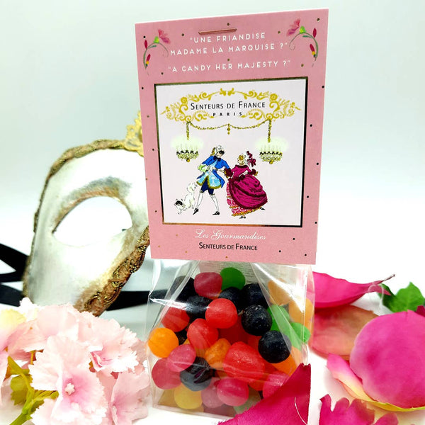 A clear bag of Senteurs De France Royal Confectionery Fruit Rolls rests on white, adorned with a pink label featuring an 18th-century couple. Pink flowers and ribbons evoke the elegance of fine French gifts by Senteurs De France.