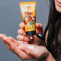 A person dispenses Barefoot Venus Apricot Brandy Hand Cream, enriched with macadamia nut oil, into their palm. The tube displays vibrant artwork of a woman and tiger on the label.
