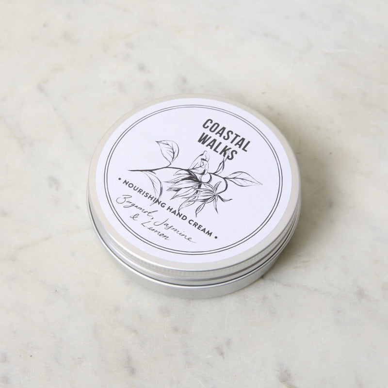 A round, white tin labeled "Norfolk Natural Living Coastal Walks Hand Cream - 100ml" sits on a light marble surface. The illustration of leaves complements the text: "Nourishing Hand Cream" with "Bergamot, Jasmine & Lemon." Infused with herbal extracts for a truly moisturizing experience.