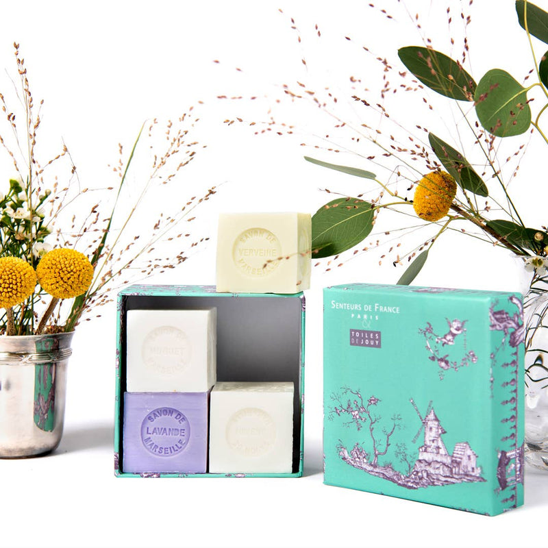 A Senteurs De France Coffret featuring Lavender, Lily of the Valley, Verbena, and Jasmine soaps is showcased in a decorative box with yellow billy ball flowers in a metal vase and sprigs of greenery nearby.