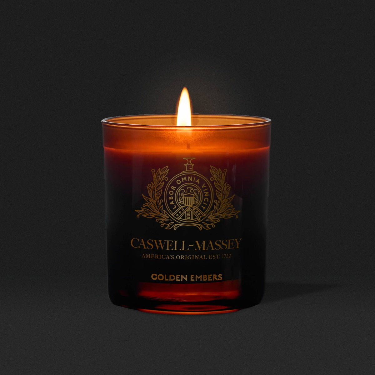 The Caswell-Massey Candle – Golden Embers glows warmly in a dark amber glass jar, its meditative fragrance and orange flame creating a cozy atmosphere.