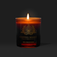 The Caswell-Massey Candle – Golden Embers glows warmly in a dark amber glass jar, its meditative fragrance and orange flame creating a cozy atmosphere.