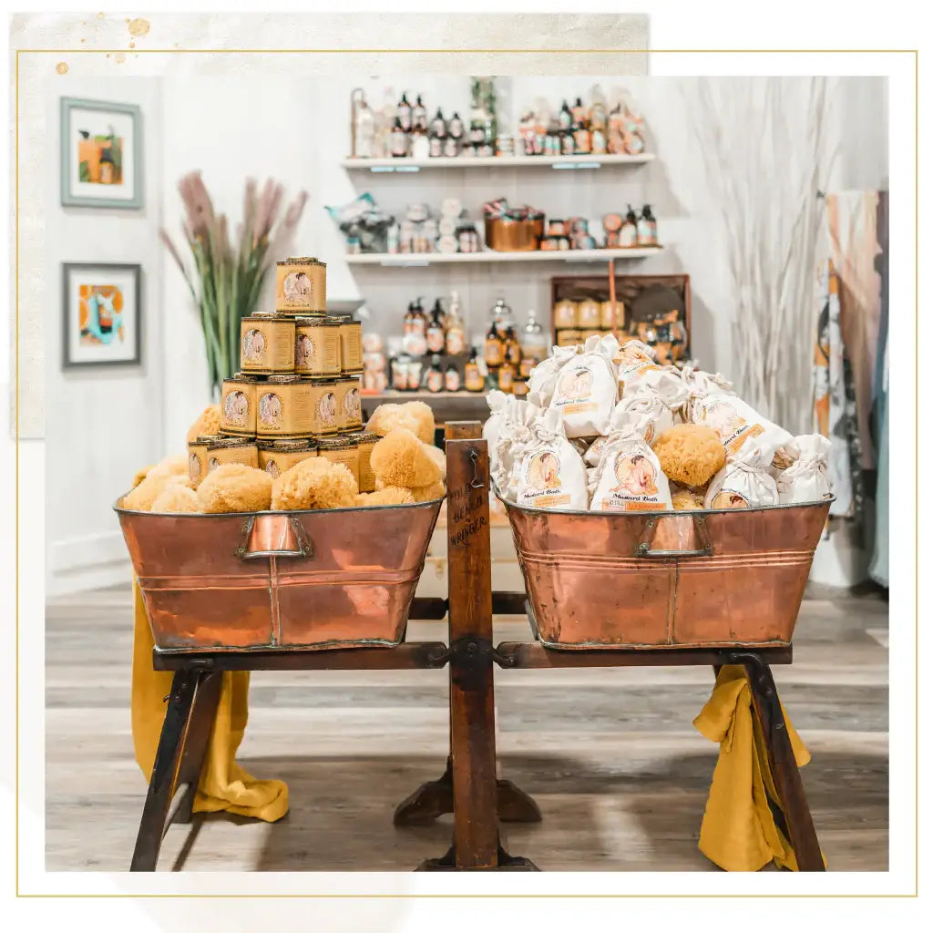 Two copper bins on a wooden stand showcase Barefoot Venus 100% Natural Mustard Bath Soak Refill Bags. Behind them, shelves display jars, plant-based formulas, and honey-themed items, all accented by framed art and decorative plants.