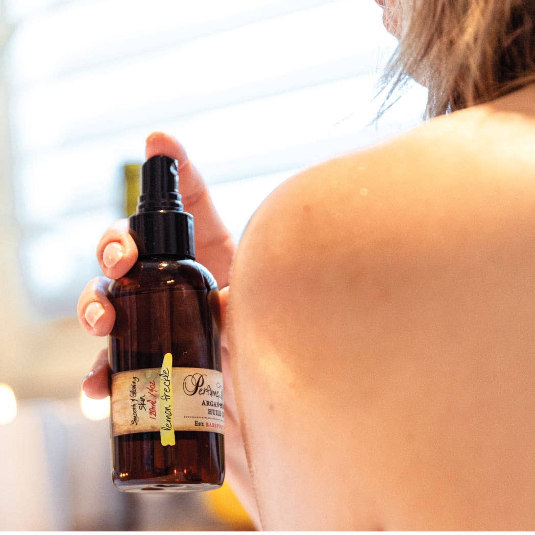 A person holds a brown spray bottle labeled "Barefoot Venus" near their bare shoulder, promising intense hydration with its plant-based formula of Lemon Freckle Argan Body Oil. The background is softly blurred, creating a warm and intimate atmosphere.