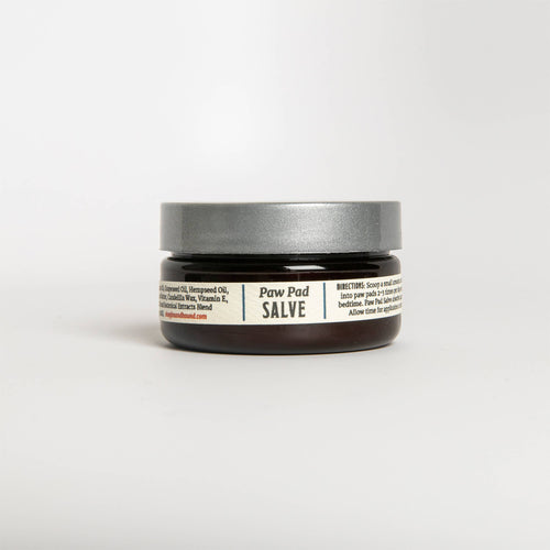 The Fox + Hound - Paw Pad Salve is a small, dark brown jar with a gray lid, promising to soothe pet paw pads with nourishing moisture through healing ingredients like beeswax and Vitamin E. The label includes usage directions for effective care.