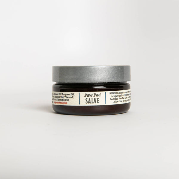 The Fox + Hound - Paw Pad Salve is a small, dark brown jar with a gray lid, promising to soothe pet paw pads with nourishing moisture through healing ingredients like beeswax and Vitamin E. The label includes usage directions for effective care.