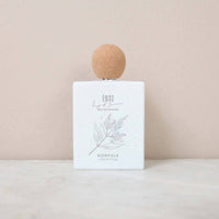 A minimalist perfume bottle with a round wooden cap rests on a light surface against a soft beige background. Its white label reads "Norfolk Natural Living 93 Days of Summer Parfum," features delicate botanical artwork, and is inspired by Norfolk Natural Living.
