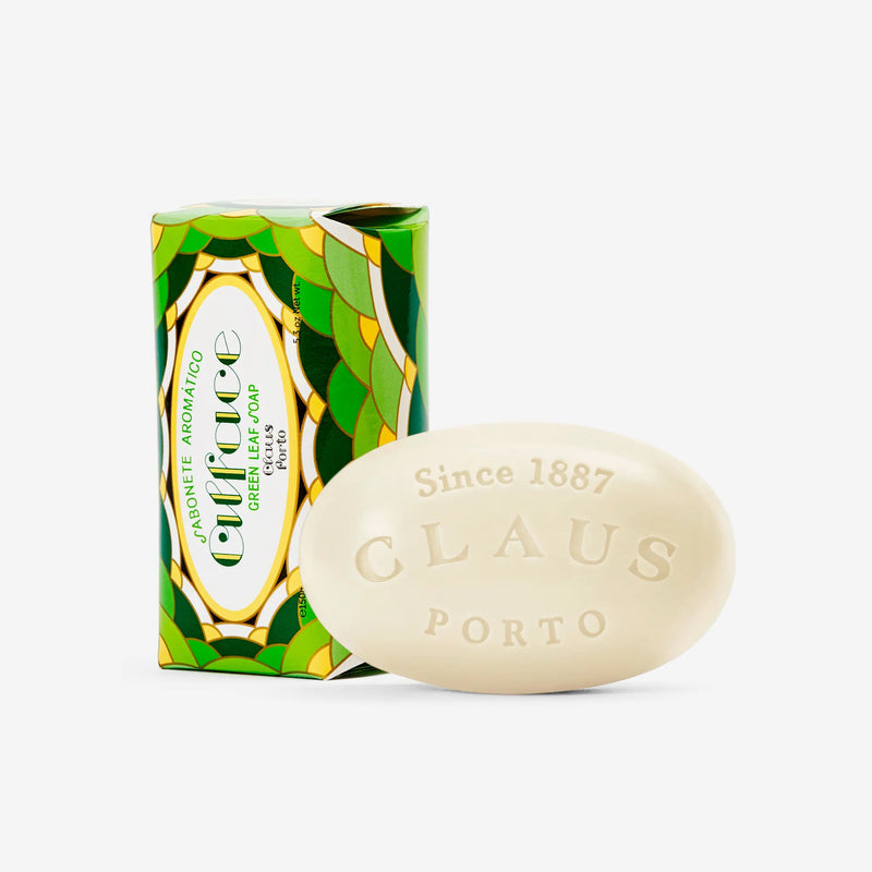 A Claus Porto 1887 Alface Green Leaf Soap bar next to its vibrant green and yellow packaging with leaf patterns. The soap is oval, white, and embossed with "Claus Porto 1887".