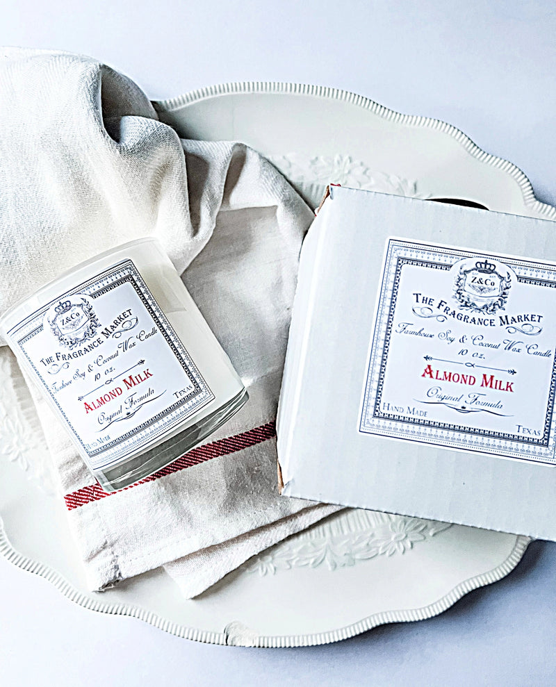 The Z&Co. Almond Milk Farmhouse Candle, in a glass jar beside its matching box, is styled on a white plate with a cream and red-striped cloth, creating a cozy, non-toxic ambiance in soft lighting.