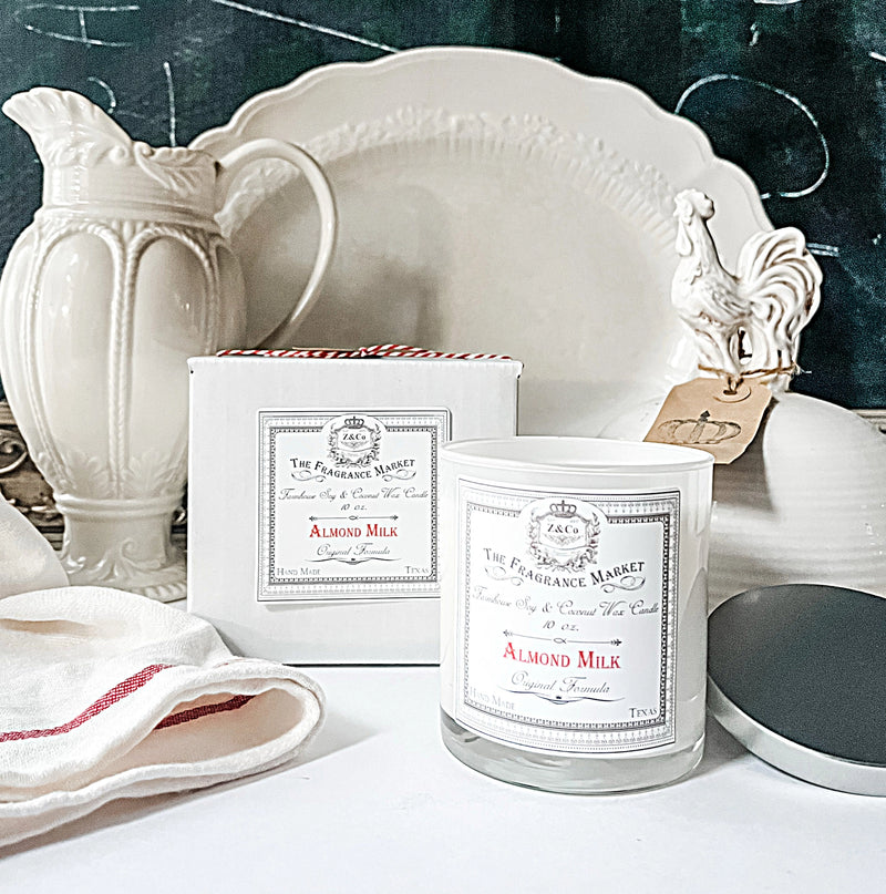 The Z&Co. Almond Milk Farmhouse Candle and its box sit on a counter with vintage ceramic dishes, a rooster figurine, a lidded pitcher, and a folded towel, creating a cozy farmhouse vibe.