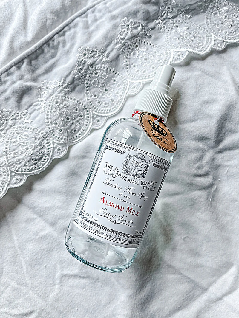 A bottle of Z&Co. Almond Milk Farmhouse Room/Linen Spray lies on a textured white fabric with lace details. The bottle features a vintage-style label and wooden cap, handcrafted in Mcallen, Texas.
