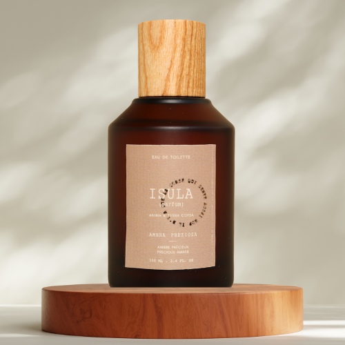 A dark amber glass bottle with a wooden cap sits on a round wooden platform. Its beige label reads "ISULA parfums Ambra Preziosa - Precious Amber eau de toilette." Soft light patterns appear in the background.