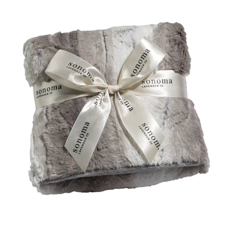 The Sonoma Lavender Angora Platinum Heat Wrap is a plush grey and white faux fur blanket, rolled up and tied with a cream-colored ribbon featuring the "Sonoma Lavender" brand name multiple times. Infused with lavender flowers, this cozy and luxurious wrap promises comfort, warmth, and soothing heat therapy.