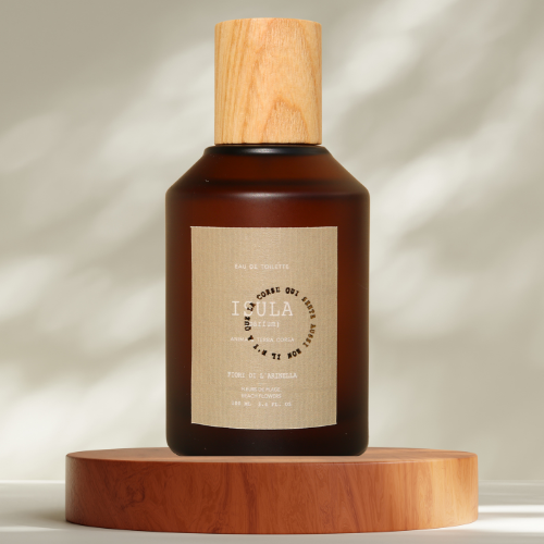 A brown glass ISULA parfums Fiore di l’Arinella - Beach Flower eau de toilette bottle with a wooden cap rests on a round wood platform. The dual-language label and gentle lighting evoke the fragrance's tranquil, beach-inspired mood.