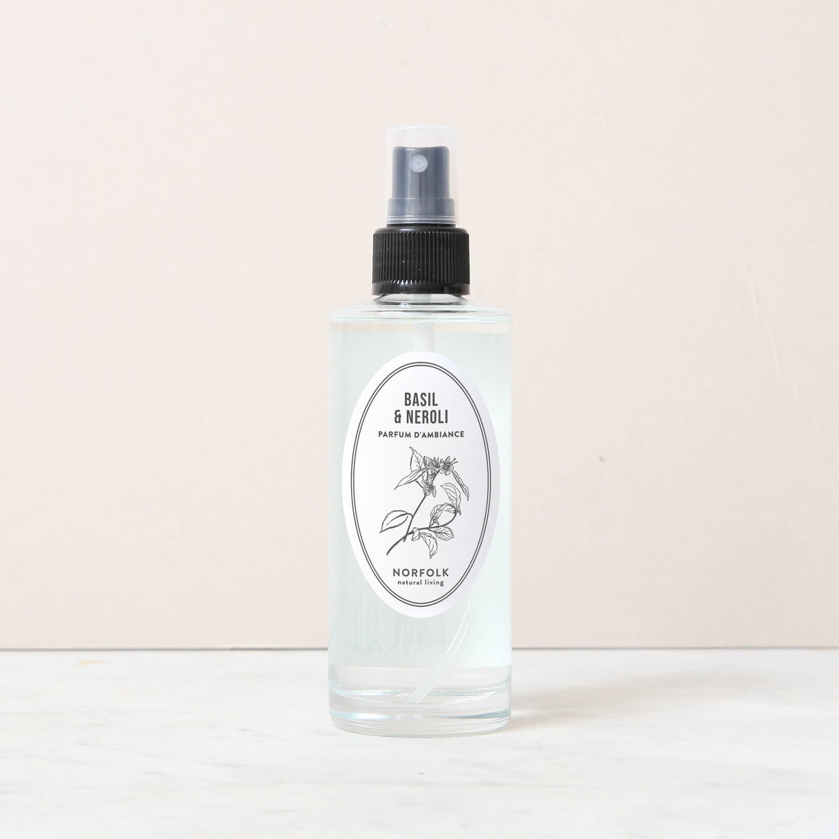 A clear 150ml spray bottle of Norfolk Natural Living Basil & Neroli Room and Mood Mist stands on a light marble surface, featuring a black nozzle and botanical label. This flame-free mist offers a Mediterranean-inspired fragrance.