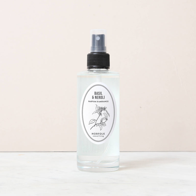 A clear 150ml spray bottle of Norfolk Natural Living Basil & Neroli Room and Mood Mist stands on a light marble surface, featuring a black nozzle and botanical label. This flame-free mist offers a Mediterranean-inspired fragrance.