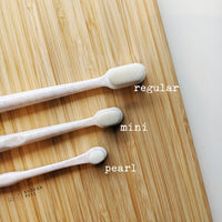 Three white Wag & Bright Supply Co. - Puppy Polisher Eco Toothbrushes, available in regular, mini, and pearl sizes, are crafted from wheat straw bioplastic and neatly arranged on a wooden surface. Each toothbrush head is labeled with its respective size, highlighting a dedication to pet dental health.