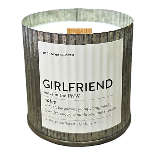 The Anchored Northwest Girlfriend soy candle features a wood wick in a rustic, ribbed metal container. With fragrance notes of powder, bergamot, ylang ylang, vanilla, lavender, sugar, sandalwood, musk, and amber; it's eco-friendly and labeled "GIRLFRIEND" made in the PNW.