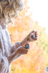 A person with wavy blonde hair, wearing a fuzzy cream-colored sweater, is holding a lighter and Barefoot Venus Coconut Kiss Roll on Perfume in their hands. The background features vibrant autumn foliage with yellow and orange leaves.