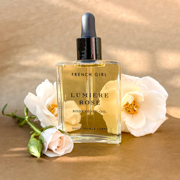 A glass dropper bottle of FRENCH GIRL Organic Hydrating Rose Body Oil (2 oz) is shown on a brown background, surrounded by white and pale pink flowers. The bottle contains light golden, hydrating oil.