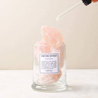 A hand holds a dropper over a glass with pink rock, highlighting the Norfolk Natural Living Rose Quartz Gemstone Diffuser—ideal for aromatherapy and emotional balance.