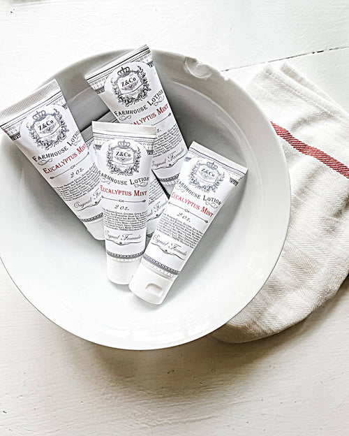 A white bowl holding several tubes of Z&Co. Eucalyptus Mint and Lavender Farmhouse Lotion gleams on a white wooden surface. This travel-sized collection from Z&Co. adds an element of luxury for those on the move. Next to the bowl lies a white cloth with a red stripe, creating a charming touch.