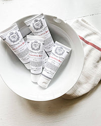 A white ceramic bowl holds five travel-sized tubes of Z&Co. Eucalyptus Mint Farmhouse Lotion in 2 oz sizes. The predominantly white tubes, featuring elegant black and red detailing, are arranged with a white cloth accented with a red stripe beside the bowl.