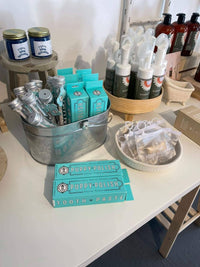 A display of pet grooming products on a table features blue boxes labeled "Puppy Polish Toothpaste" alongside tins in a metal bucket and spray bottles. Enhancing pet dental health, the exhibit includes jars on a tiered stand, pouches in a woven basket, and Wag & Bright Supply Co.'s Puppy Polisher Eco Toothbrushes (Medium).