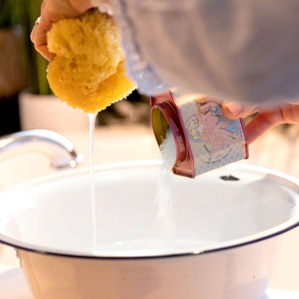 A person squeezes a yellow sponge over a white basin while pouring Barefoot Venus Coconut Kiss Bath Soak from Barefoot Venus from a metal container. The cleaning solution is being prepared at the kitchen sink area.