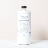 A white bottle of Norfolk Natural Living Alternative Bleach, an eco-friendly disinfectant with a black cap and minimalist label, stands upright on a light surface against a pale background, reflecting its non-toxic cleaner formula.
