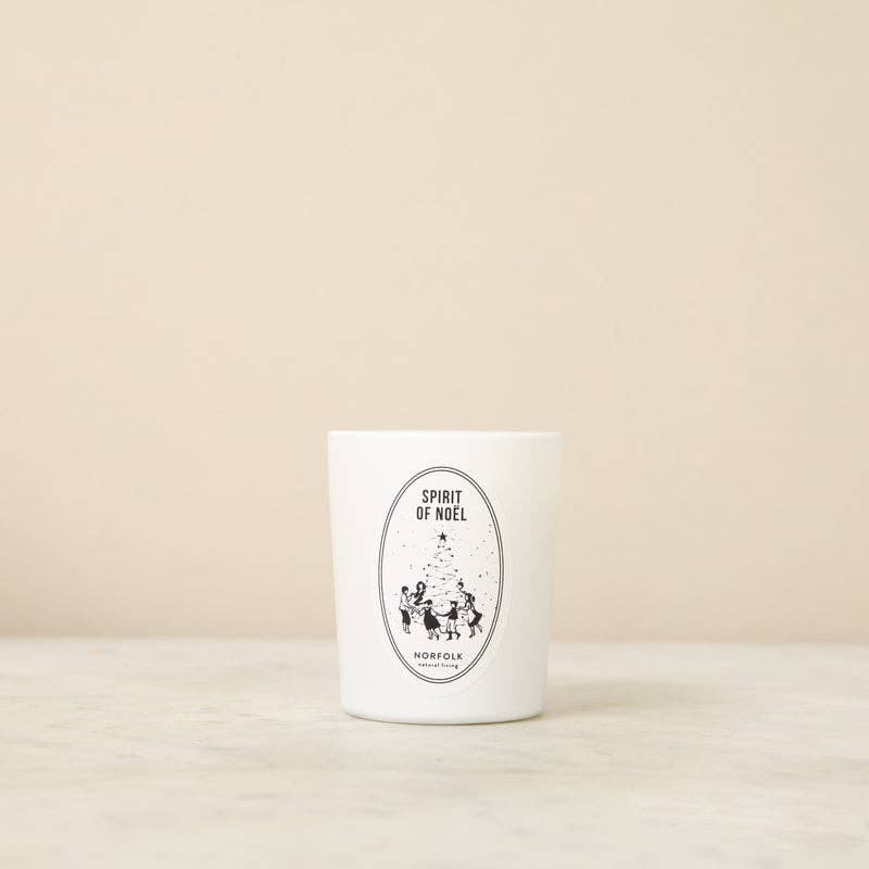 The Norfolk Natural Living Spirit of Noel Christmas Votive Candle sits on a light surface with a “Spirit of Noel” label depicting people by a Christmas tree. It’s white and scented with orange blossom balsam.